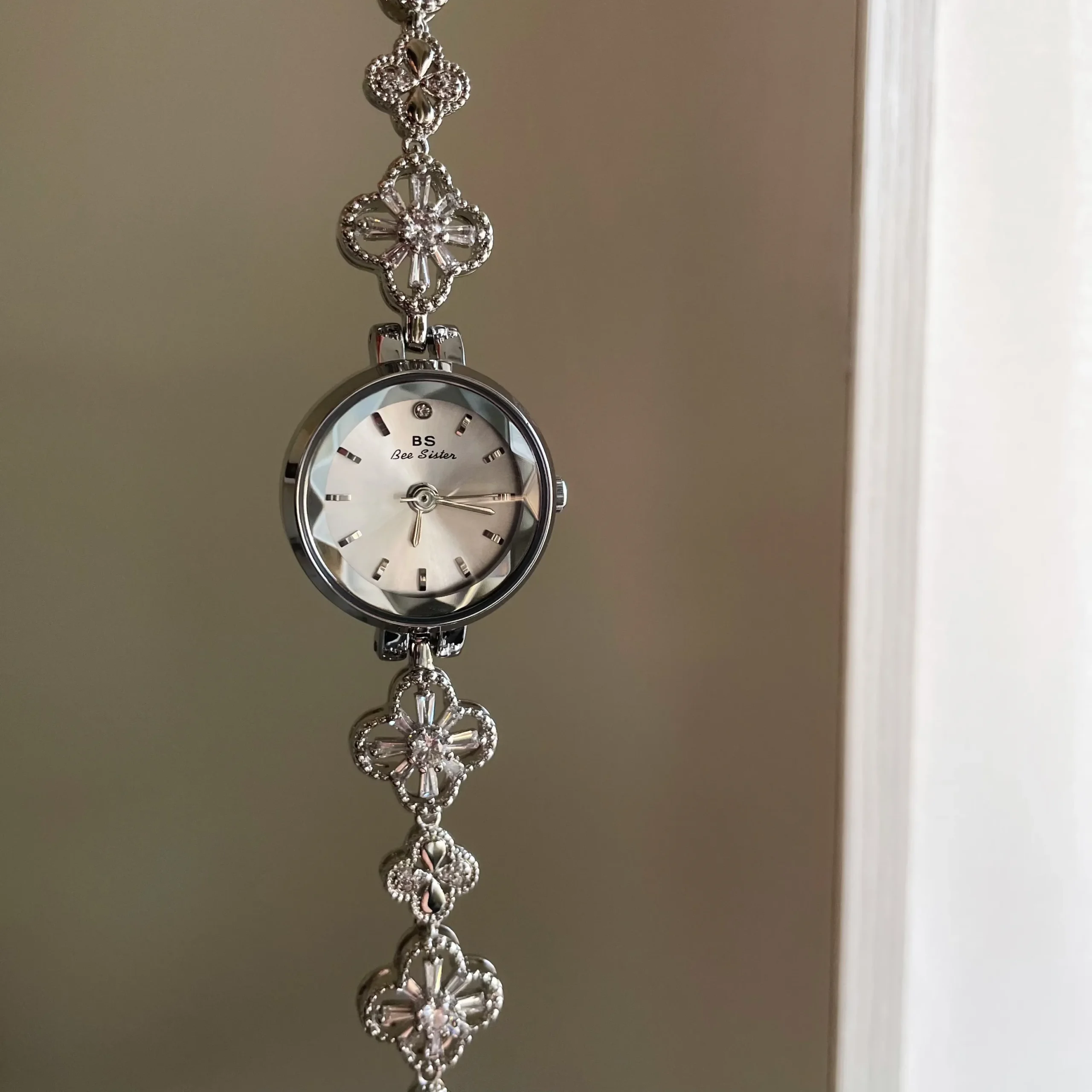 A silver clover-link bracelet watch suspended against a soft background, clearly showing the faceted crystal and mother-of-pearl dial with the BS Bee Sister logo.