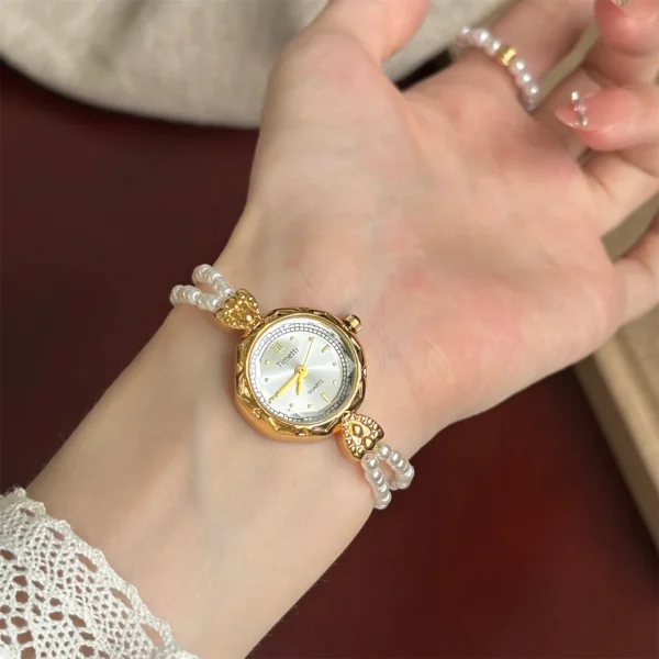 Close-up of a hand wearing a vintage gold watch with a delicate double-strand pearl bracelet. The elegant timepiece is displayed against a white lace cuff.