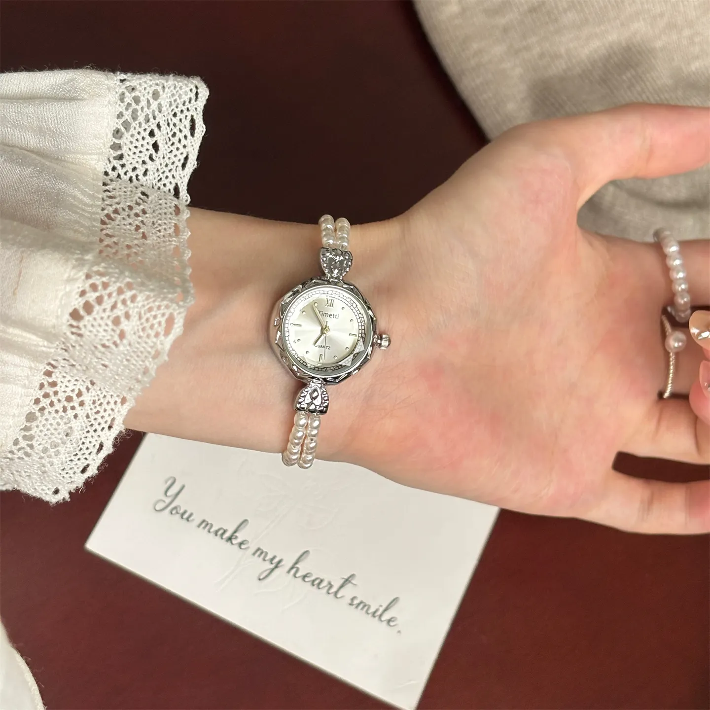 Elegant silver-toned watch worn on a wrist, featuring a quartz dial and a bracelet made of double-strand small white pearls, next to a card that says "You make my heart smile."